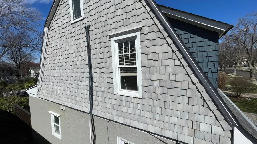 costly window mistake repairs Side view of a house with grey shingle siding, two windows, and a blue side wall, under a clear blue sky.